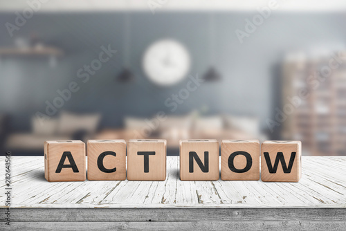 Fotografiet  Act now message sign on a wooden desk
