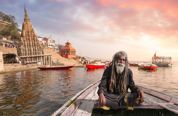 Indian sadhu baba on a wooden boat overlooking ancient Varanasi city architecture with Ganges river ghat at sunset