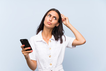 young woman with a mobile phone over isolated blue wall having doubts and with confuse face expressi
