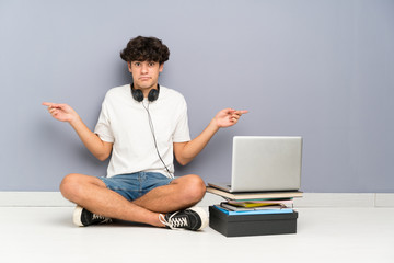 Young man with his laptop sitting one the floor pointing to the laterals having doubts