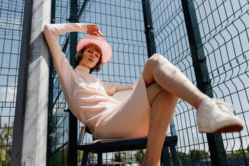 Stylish girl in a gently pink dress and pink visor sitting on a chair behind fence of the lattice at the sports field on a summer sunny day