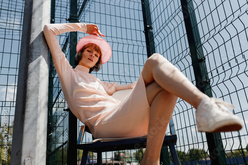 Stylish girl in a gently pink dress and pink visor sitting on a chair behind fence of the lattice at the sports field on a summer sunny day