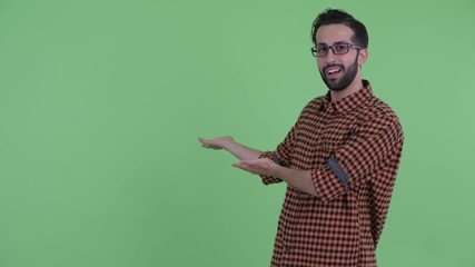 Wall Mural - Happy young bearded Persian hipster man talking while showing to back