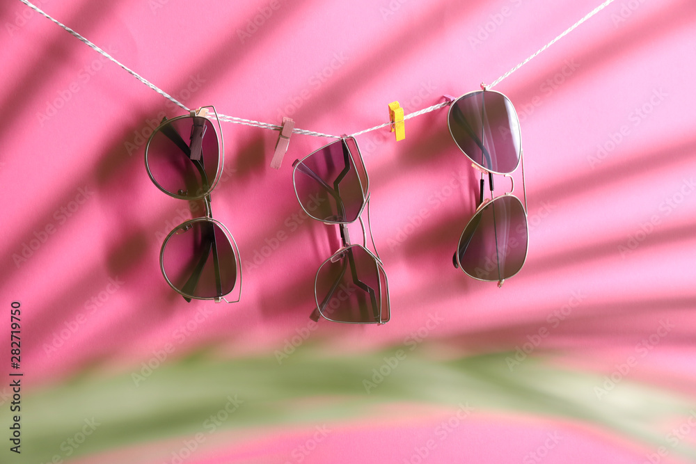 Stylish sunglasses hanging on rope against color background, view through tropical leaf