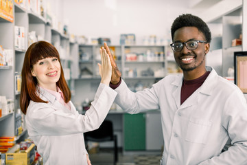 Wall Mural - Two happy colleages pharmacists, African man and Caucasian woman working in drugstore, smiling. giving five and having fun.