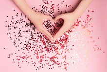 Beautiful Female Hands With Vibrant Red Sparkling Heart-shaped Confetti Poured On Pink Background. Flat Lay. Love And Celebration Concept.