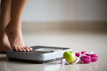 Female Leg Stepping On Weigh Scales With Measuring Tape, Pink Dumbbell And Green Apple. Healthy Lifestyle, Food And Sport Concept.
