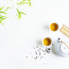 Asian tea concept, two white cups of tea and teapot surrounded with green tea dry leaves view from above, space for a text on white background.