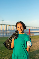 Beautiful volunteer woman posing in city park. Happy young Latin woman standing on grass, holding rake and plastic bag, smiling at camera Volunteer portrait concept