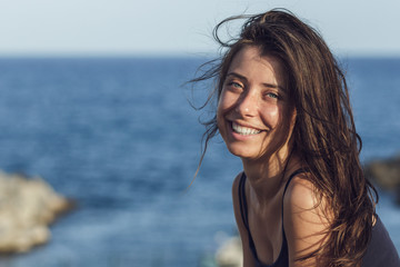 Close-up portrait of cheerful suntanned young woman looking into camera over sea and blue sky background with copy space. Photo of beautiful brunette girl relaxing on the resort under the hot sun.