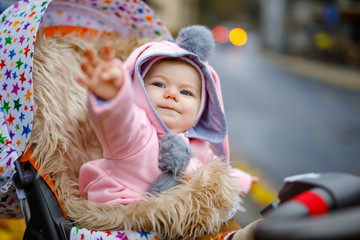 Canvas Print - Cute little beautiful baby girl sitting in the pram or stroller on autumn day. Happy smiling child in warm clothes, fashion stylish pink baby coat with bunny ears. Baby going on a walk with parents.