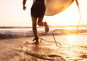 Wall Mural - Young man Surfer taking surfboard and running with long surf board to waves on the evening sunset sky background. Active holidays spending concept.