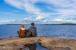 © Tatyana_Andreyeva - Tourists man and woman sitting on northern lake shore in summer day. People relaxing and admiring beautiful landscape. Travelling and discovering distant places of Earth. Onega lake, Karelia, Russia