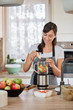 © nenadaksic - Beautiful Caucasian smiling woman in apron putting vegetables and fruits in blender while standing in kitchen.