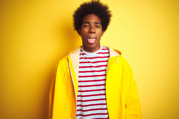 African american man with afro hair wearing rain coat standing over isolated yellow background sticking tongue out happy with funny expression. Emotion concept.