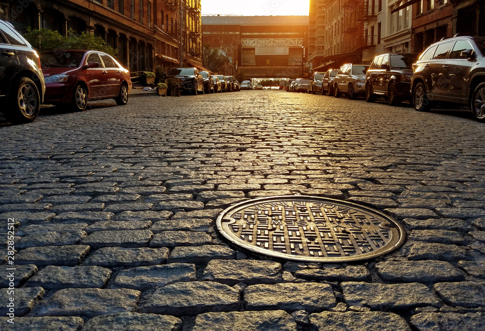 Sunlight shining on a cobblestone street and manhole cover in New York City - obrazy, fototapety, plakaty 