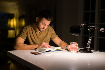 Young handsome man studying at home, reading a book at night