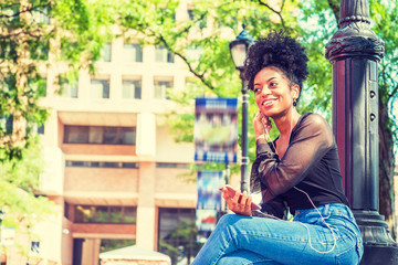 Wall Mural - Young African American Woman with afro hairstyle wearing mesh sheer long sleeve shirt blouse, blue jeans, sitting by light pole on campus in New York City, listening music with earphone and cell phone