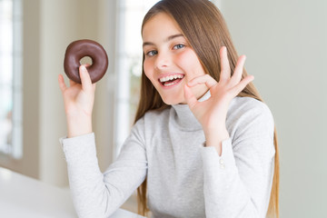 Beautiful young girl wearing eating a chocolate donut doing ok sign with fingers, excellent symbol