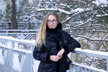 A cute, young, beautiful girl in a blue warm coat with a hood decorated with fur walks in a snowy, winter park.