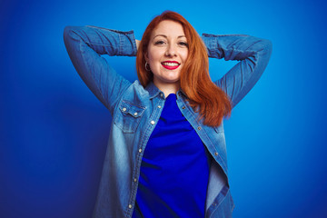 Sticker - Young beautiful redhead woman wearing denim shirt standing over blue isolated background relaxing and stretching, arms and hands behind head and neck smiling happy