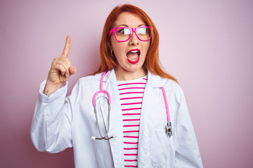 Sticker - Young redhead doctor woman using stethoscope standing over isolated pink background pointing finger up with successful idea. Exited and happy. Number one.