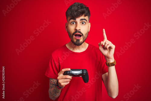 Fotografía Young gamer man with tattoo playing video game standing over isolated red backgr