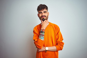Wall Mural - Young man with tattoo wearing orange sweater standing over isolated white background looking confident at the camera smiling with crossed arms and hand raised on chin. Thinking positive.