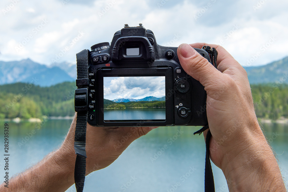 Hands of a male photographer holding a digital camera taking pictures of a idyllic landscape with a lake and mountains while the picture shows at the display - obrazy, fototapety, plakaty 