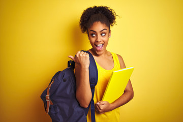 Wall Mural - American student woman wearing backpack holding notebook over isolated yellow background pointing and showing with thumb up to the side with happy face smiling