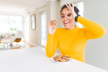 Young african american girl eating chocolate chips cookies as sweet snack with happy face smiling doing ok sign with hand on eye looking through fingers