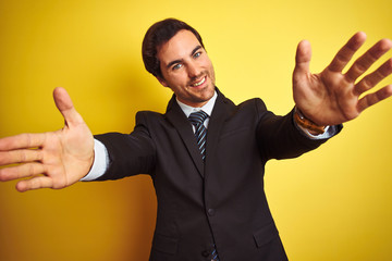 Young handsome businessman wearing suit and tie standing over isolated yellow background looking at the camera smiling with open arms for hug. Cheerful expression embracing happiness.