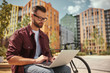 © Friends Stock - I can work from anywhere. Young handsome man with stubble in casual clothes and eyeglasses working on laptop while sitting on the bench near his bicycle