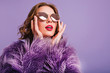 © Look! - Close-up photo of inspired trendy lady in sparkle glasses looking up with mouth open. Indoor portrait of elegant young woman in purple fur coat.