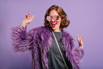 Wall Mural - Stunning cute girl with curly brown hair posing with pleasure on purple background. Indoor photo of dancing young lady in trendy fur coat laughing to camera.