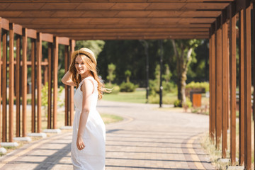 beautiful girl in white dress touching straw hat while walking near wooden construction and looking at camera