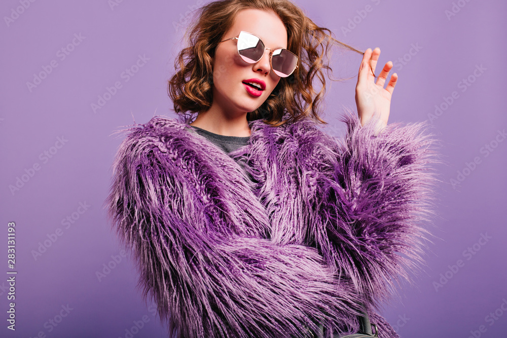 Dreamy pretty girl plays with short curly hair standing on bright background. Indoor portrait of pensive female model in sunglasses and purple fluffy coat. - obrazy, fototapety, plakaty 