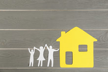 Paper Figures Of Happy Family Holding Hands And Yellow House On Grey Wooden Background, Flat Lay