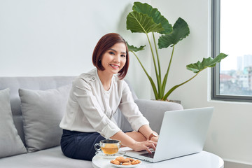 Sticker - Beautiful asian woman sitting in her living room and refreshing with cup of tea and biscuits