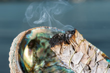 Smudging Ritual Using Burning Thick Leafy Bundle Of White Sage In Bright Polished Rainbow Abalone Shell On The Beach At Sunrise In Front Of The Lake.