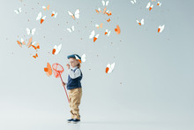Cute And Blonde Kid In Retro Vest And Cap Holding Butterfly Net On Grey Background With Fairy Butterflies