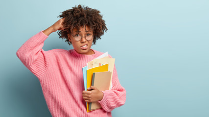 Wall Mural - Education concept. International student studies in college, holds documents and notepads, scratches head with puzzlement, wears optical glasses, pink oversized sweater, isolated on blue wall