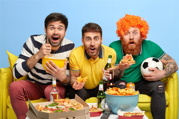 Excited three male friends concentrated at screen of TV set, watch football match with great interest, pose on sofa in spacious living room, eat popcorn, pizza, crisps, drink beer, have fun at home.