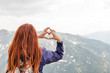Girl making a heart shape with mountain landscape in the background. Copyspace. A young pretty redheaded woman standing on a background of mountains. Trekking, vacation and tourism concept. Copyspace