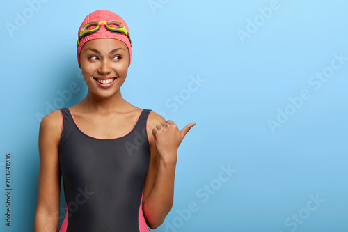 Fotografía Professional female swimmer points away on blank space, wears pink swimcap and bathing suit, attracts your attention to awesome promo, has clean skin, healthy body and slim figure