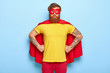Photo of serious male in superhero costume, keeps hands on waist, possesses extraordinary talents, ready to protect our universe, isolated on blue wall. Bearded guy in red mask, cape, yellow t shirt