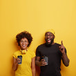 © wayhome.studio  - Happy black students have fun after lectures, drink takeaway coffee, hipster guy in yellow hat and black t shirt, points index finger above on copy space, carefree Afro woman wears headphones on neck