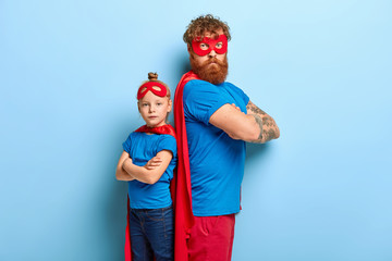 Self confident father and daughter stand back to each other, keep arms folded, look seriously at camera, wear special superhero costumes, play game, isolated on blue background. Ginger family