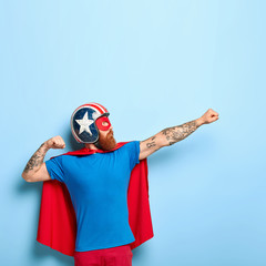 Vertical shot of bearded man makes flying gesture, clenches fists, has goal to achieve, wears protective headgear, red cape, pretends being heroic character, has superhuman power isolated on blue wall