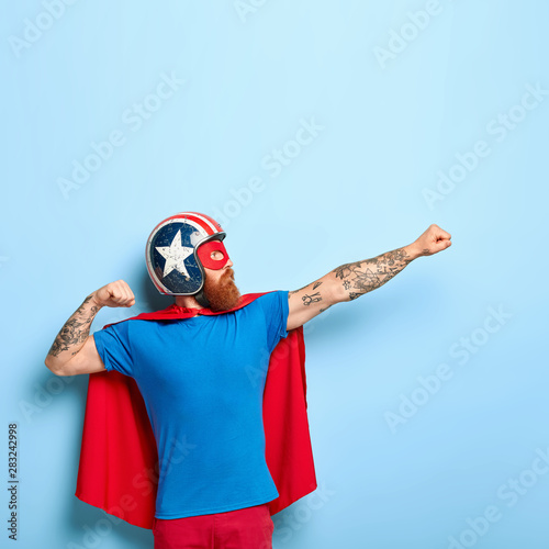 Vertical shot of bearded man makes flying gesture, clenches fists, has goal to achieve, wears protective headgear, red cape, pretends being heroic character, has superhuman power isolated on blue wall